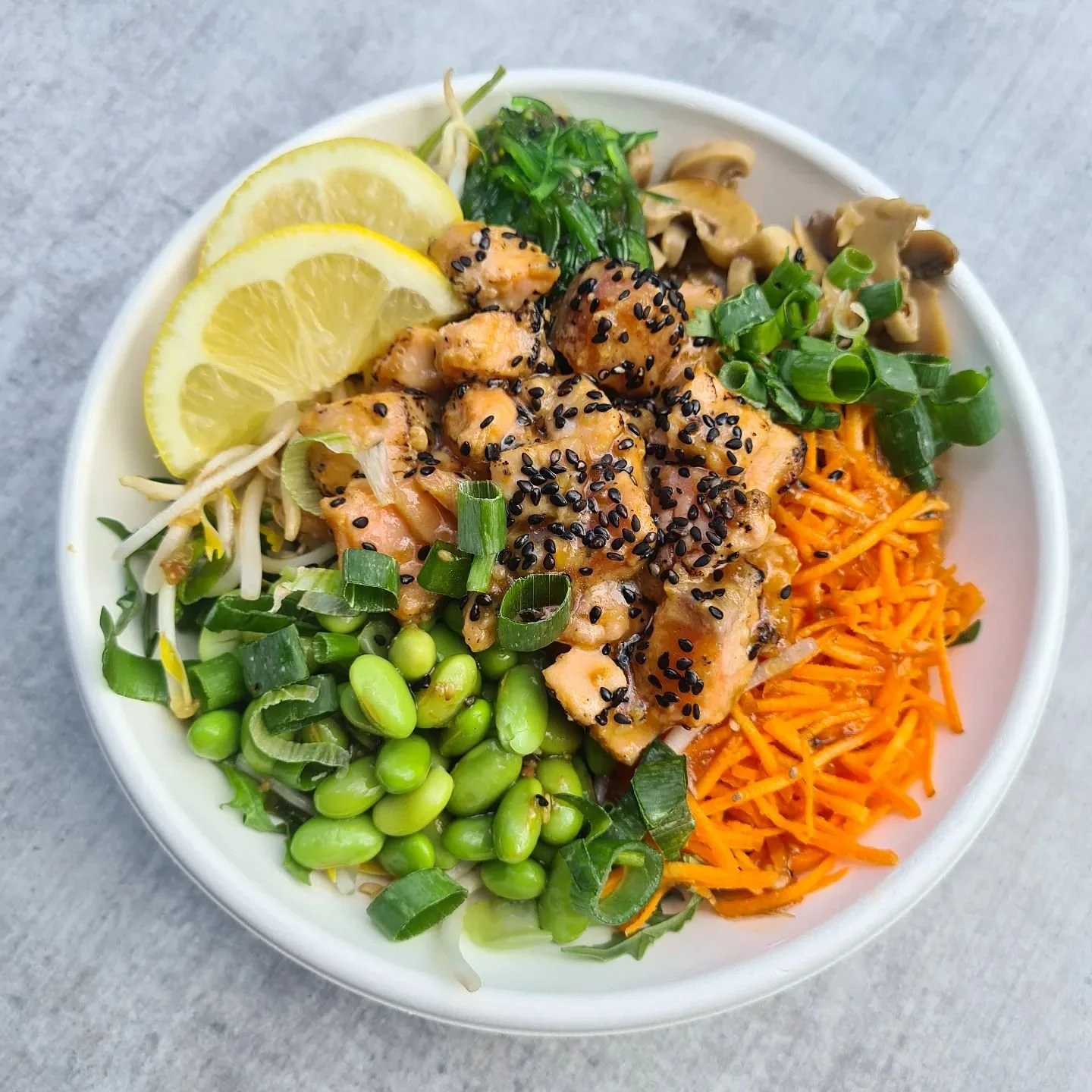 A bowl of salad with grilled chicken topped with black sesame seeds, shredded carrots, green onions, edamame, bean sprouts, sliced lemon, and seaweed.