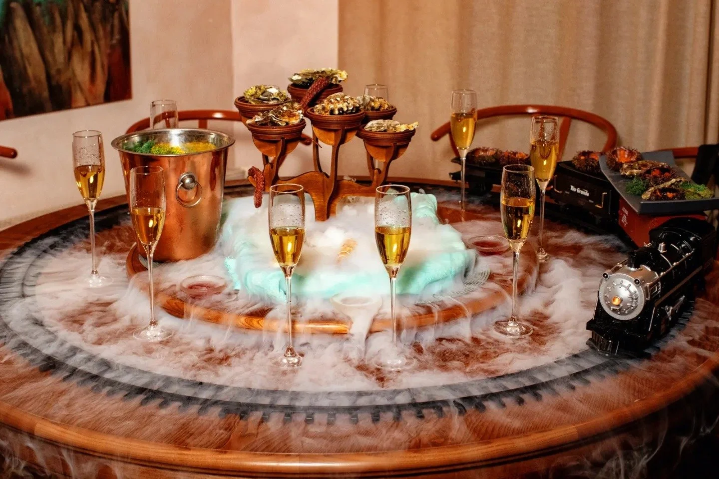 A tabletop set with a miniature train, multiple glasses of champagne, a copper ice bucket, and various food items, surrounded by smoke or fog for decoration.