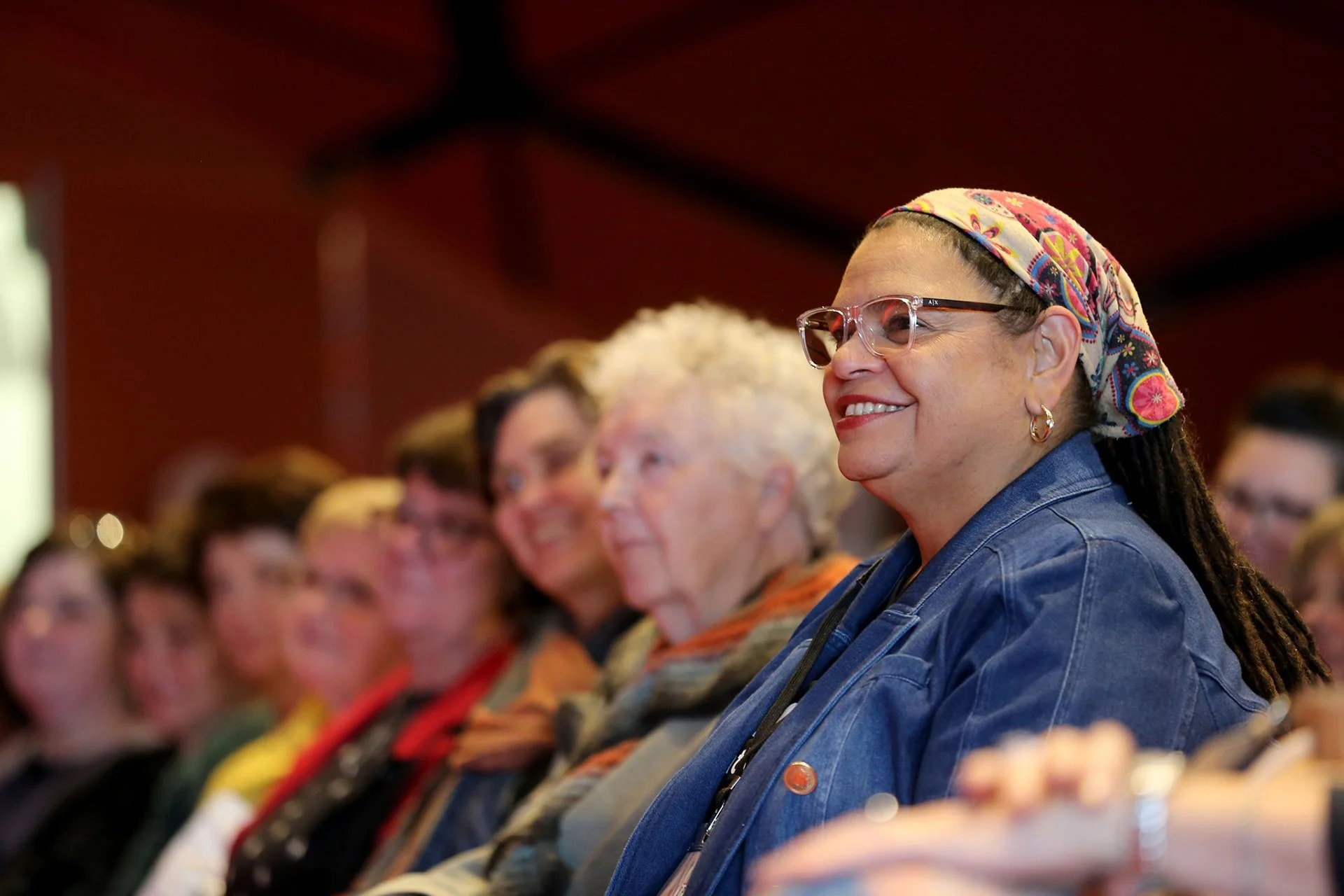 A group of women sitting in an indoor venue, smiling and listening attentively. The woman in the foreground has glasses, earrings, a colorful headscarf, and a denim jacket.