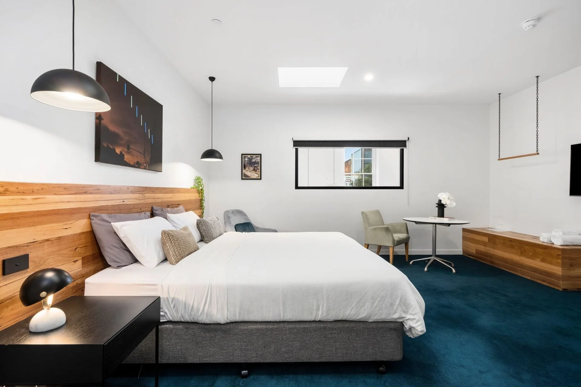 Modern bedroom with white walls, wooden headboard, and dark blue carpet. Contains a bed with white bedding, gray and beige pillows, a black bedside table with a lamp, a small sitting area with chairs, a window with a black frame, and wall art.
