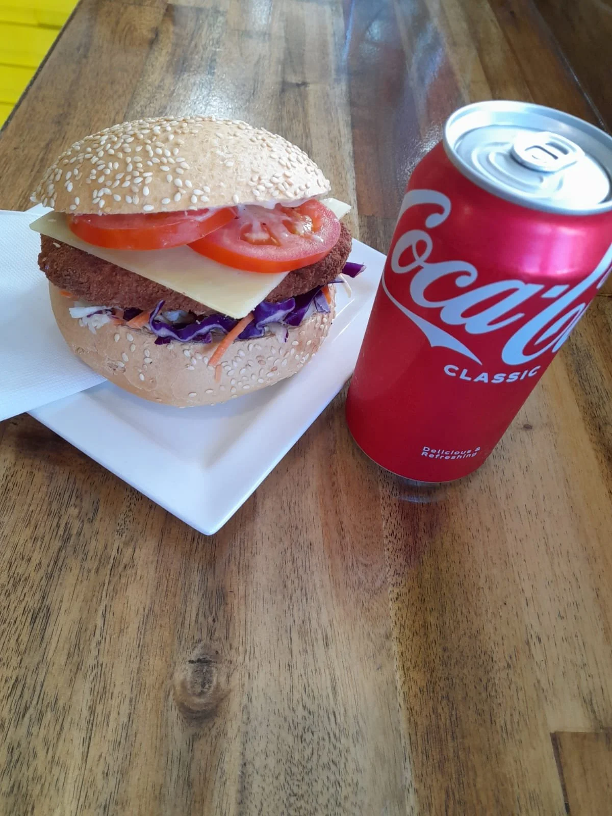 A cheeseburger with tomato slices, cheese, a breaded patty, and purple cabbage in a sesame seed bun, served on a white plate with a napkin, and a can of Coca-Cola classic on a wooden table.