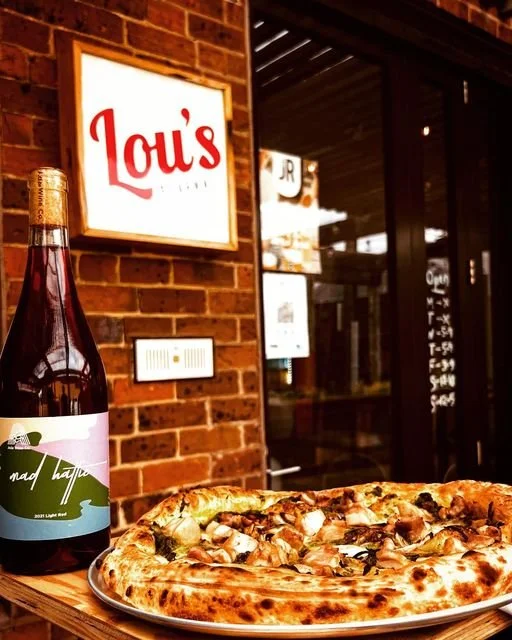 A pizza with cheese, mushrooms, and other toppings on a tray, a bottle of soda labeled 'Mad Hatter,' and a sign for Louis's on a brick wall outside a restaurant.