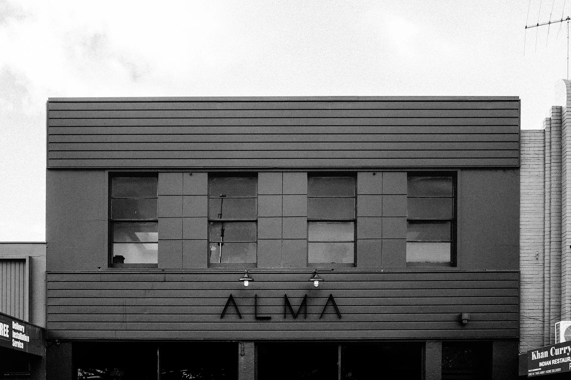Black and white photo of a modern building with the sign 'ALMA' on the front, featuring large rectangular windows and horizontal siding.