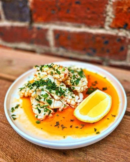 A white plate of cooked squid garnished with chopped herbs, served with lemon wedge in a flavorful sauce.