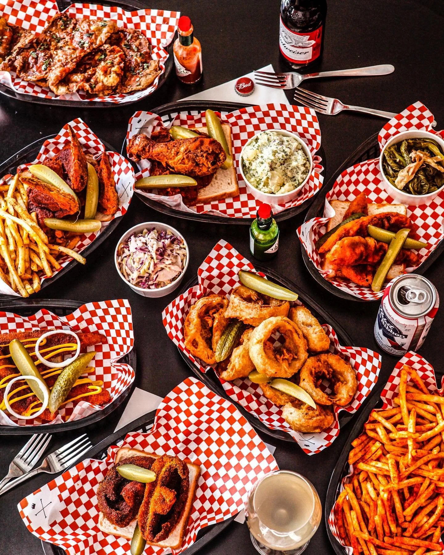 Various plates of fried chicken, onion rings, French fries, coleslaw, mashed potatoes, stewed greens, and different sauces on a black table with a Budweiser beer bottle and a can, and utensils.