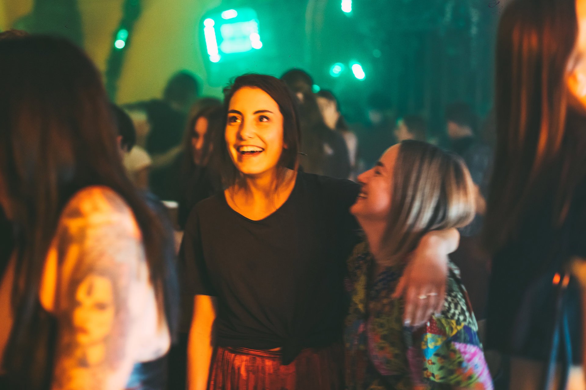 A group of women at a lively social gathering or party, smiling and engaging in conversation, with colorful lighting and a blurred crowd in the background.