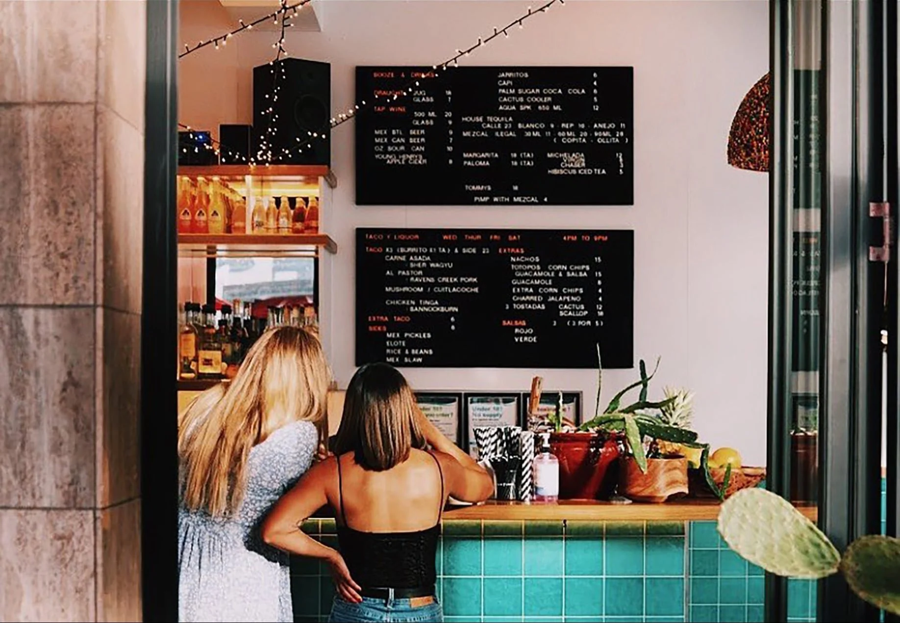 Two women are standing at a counter, looking at a menu board inside a restaurant.