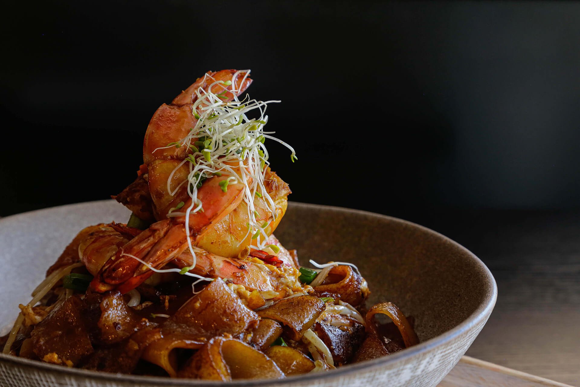 Seafood pasta dish with shrimp, potatoes, and vegetables topped with sprouts, served in a textured bowl against a dark background.