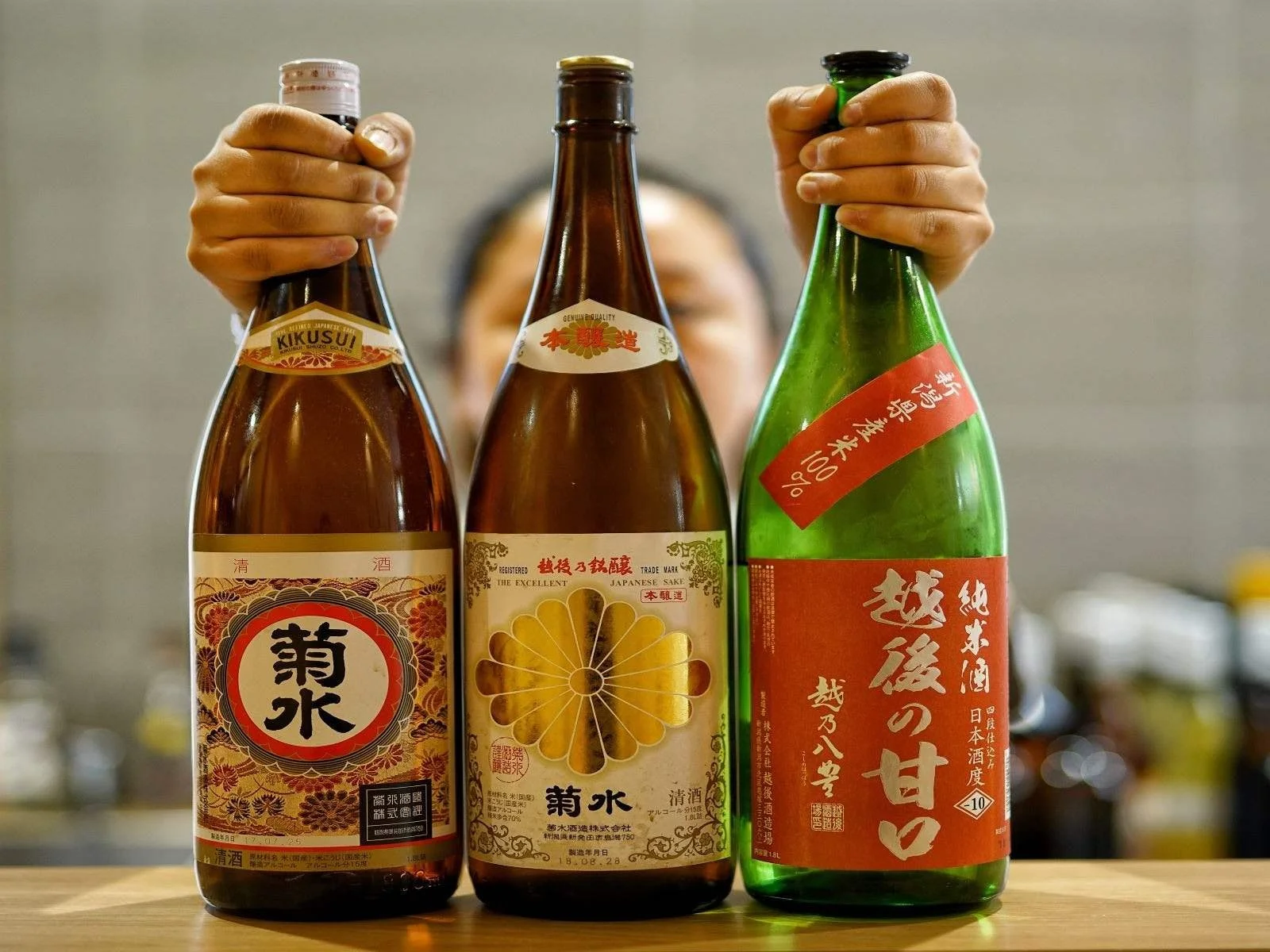 Three bottles of Japanese sake on a wooden surface, with a person's hands holding the bottles. The background is blurred.