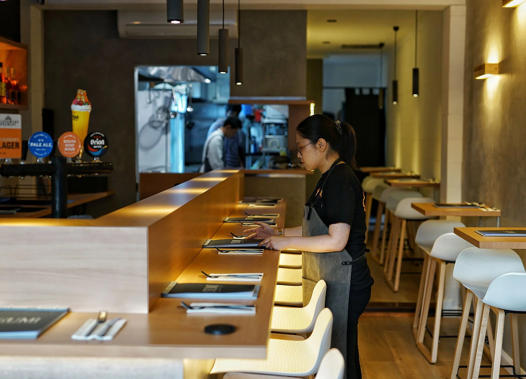 A waitress standing at a long wooden counter in a modern restaurant, looking at a menu or check, with empty chairs arranged along the bar. Two people are visible in the background, working in the kitchen area.