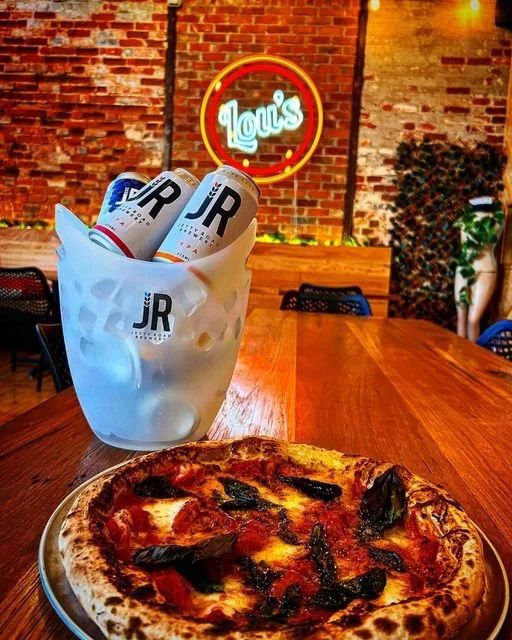 Pizza on a plate, a frosted glass containing cans of JR beer, and a wooden table with a brick wall background and a neon sign reading 'Lou's' in a restaurant setting.