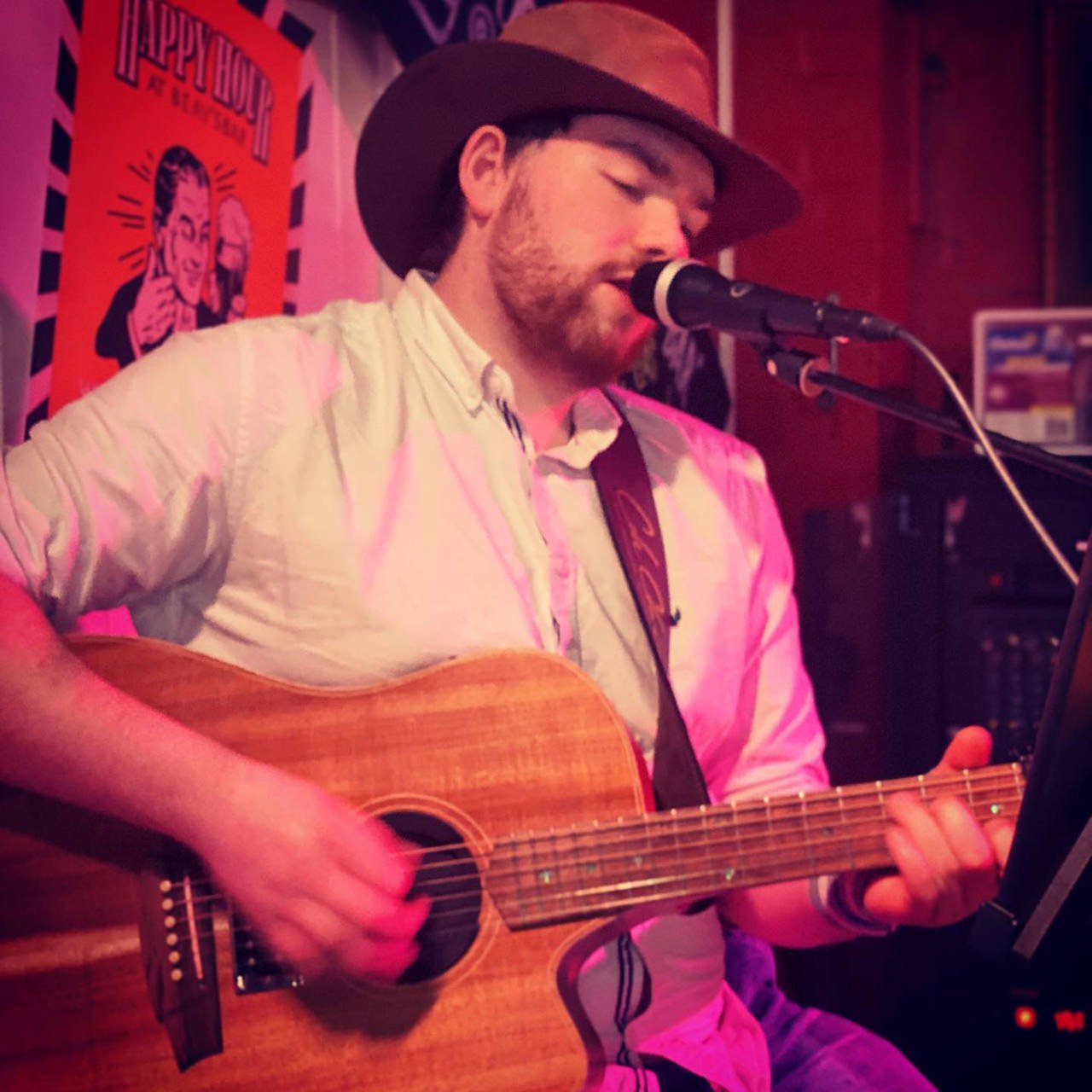 A man wearing a cowboy hat and a white shirt playing an acoustic guitar and singing into a microphone in a dimly lit venue with a colorful poster in the background.