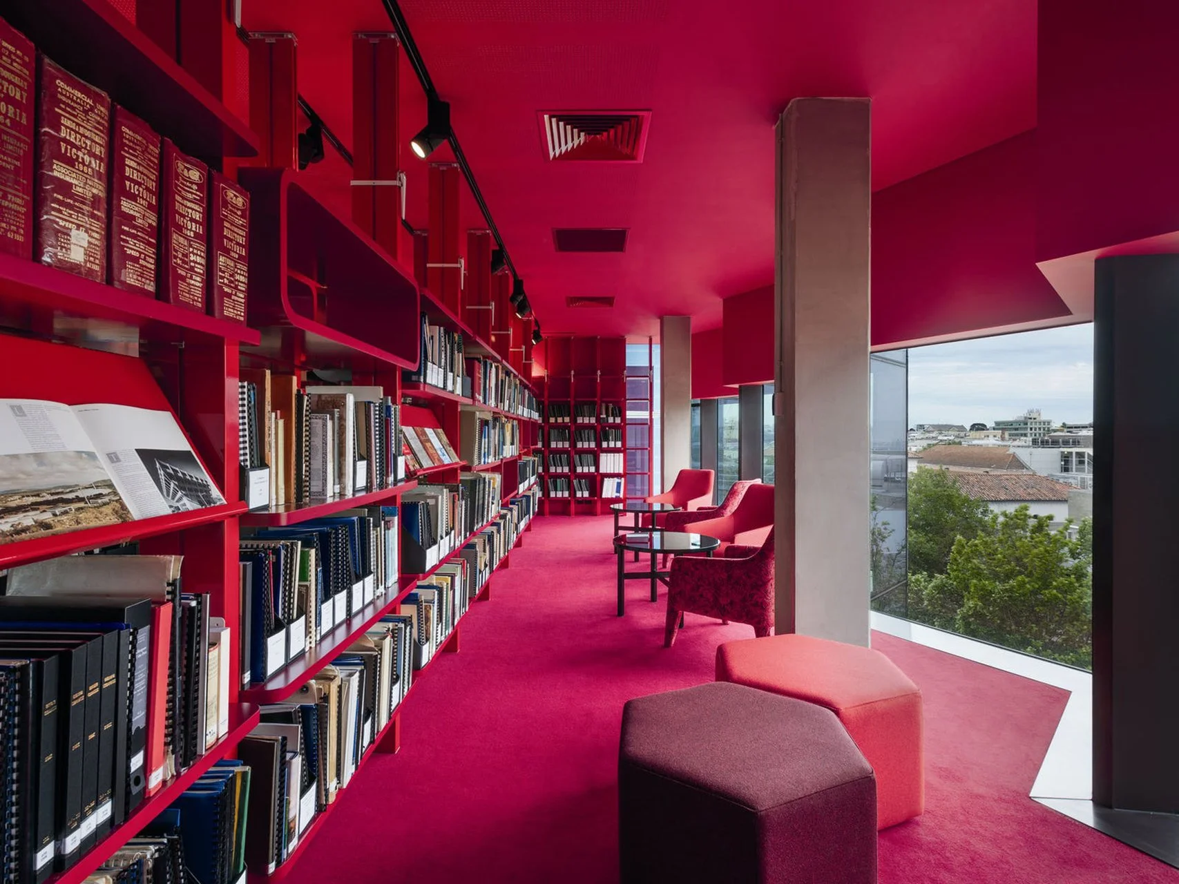 Interior of a modern library or reading area with pink walls and carpet, bookshelves filled with books, and seating including pink cushions and chairs near large windows showing a cityscape view.