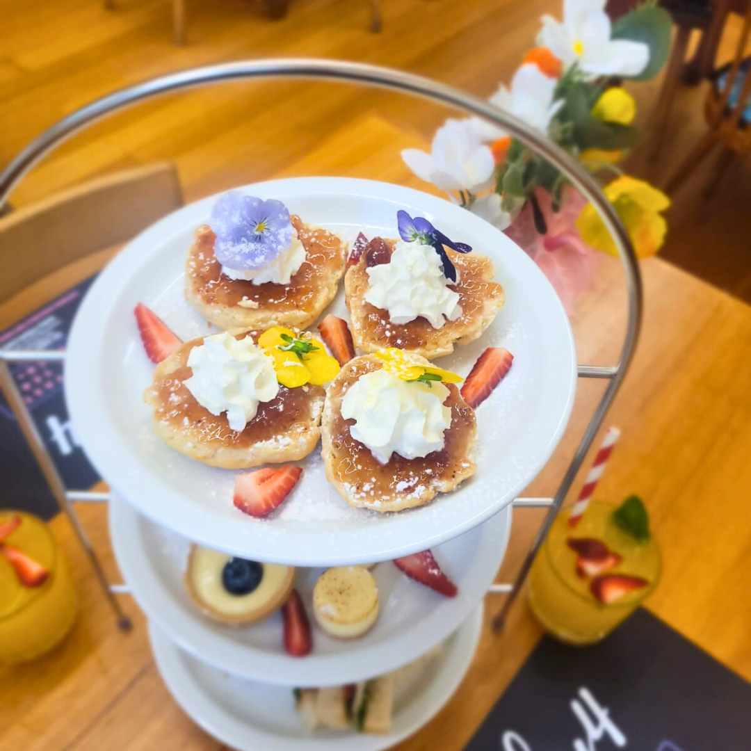 A three-tiered serving stand with small desserts on the top tier, including mini tarts topped with whipped cream and edible flowers, garnished with slices of strawberries. The lower tiers contain more desserts such as cookies and small pastries. In t