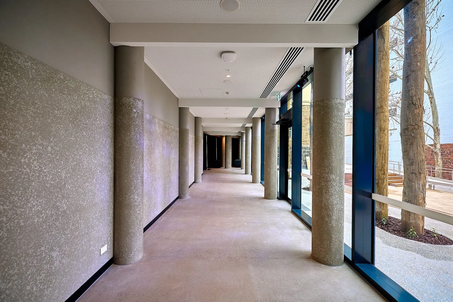 Empty hallway with large windows showing trees outside, concrete columns, beige floor and textured wall surfaces.