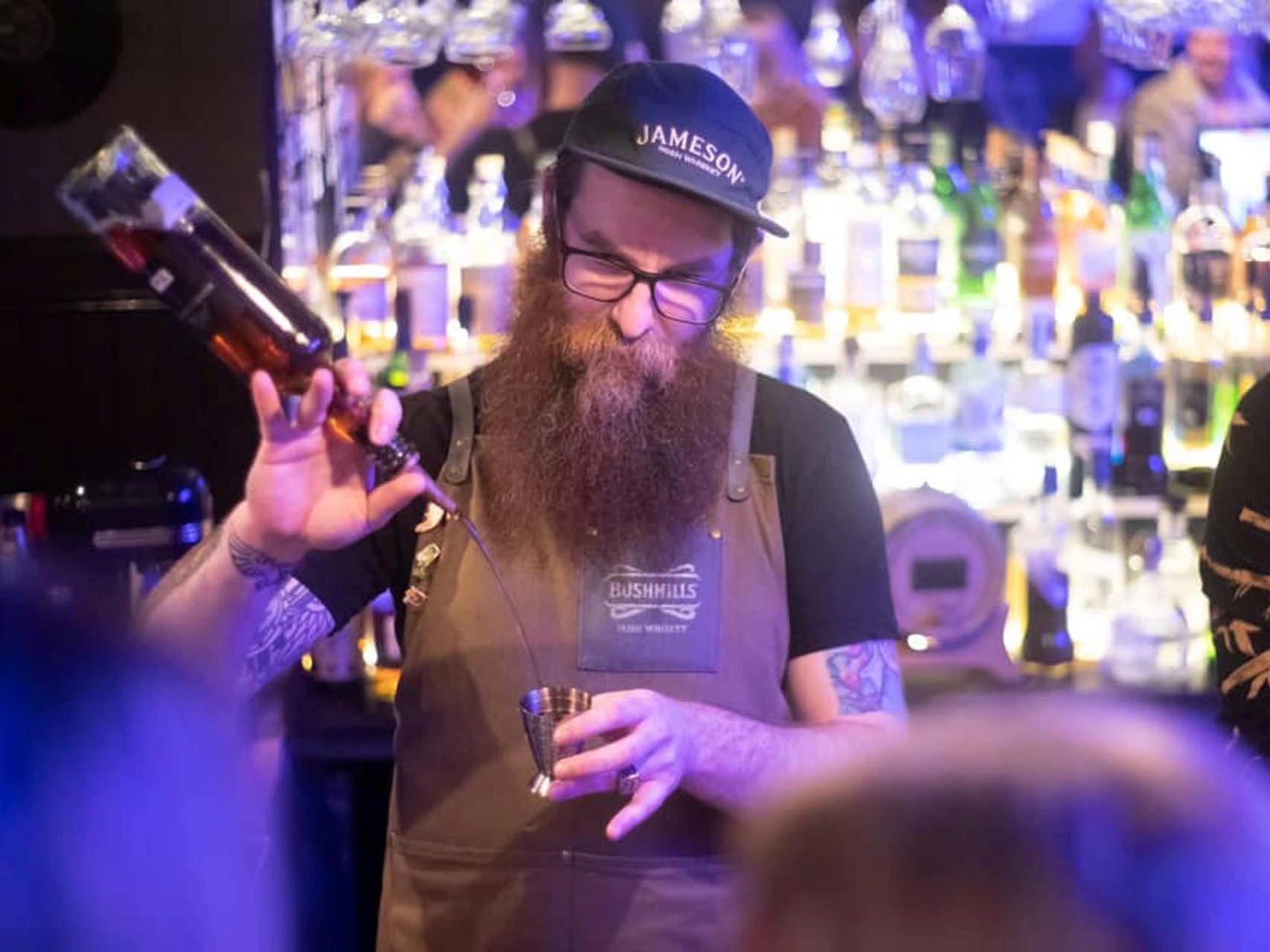 A bearded man with glasses pouring a drink from a bottle into a metal jigger at a bar, with a colorful liquor bottle display in the background.