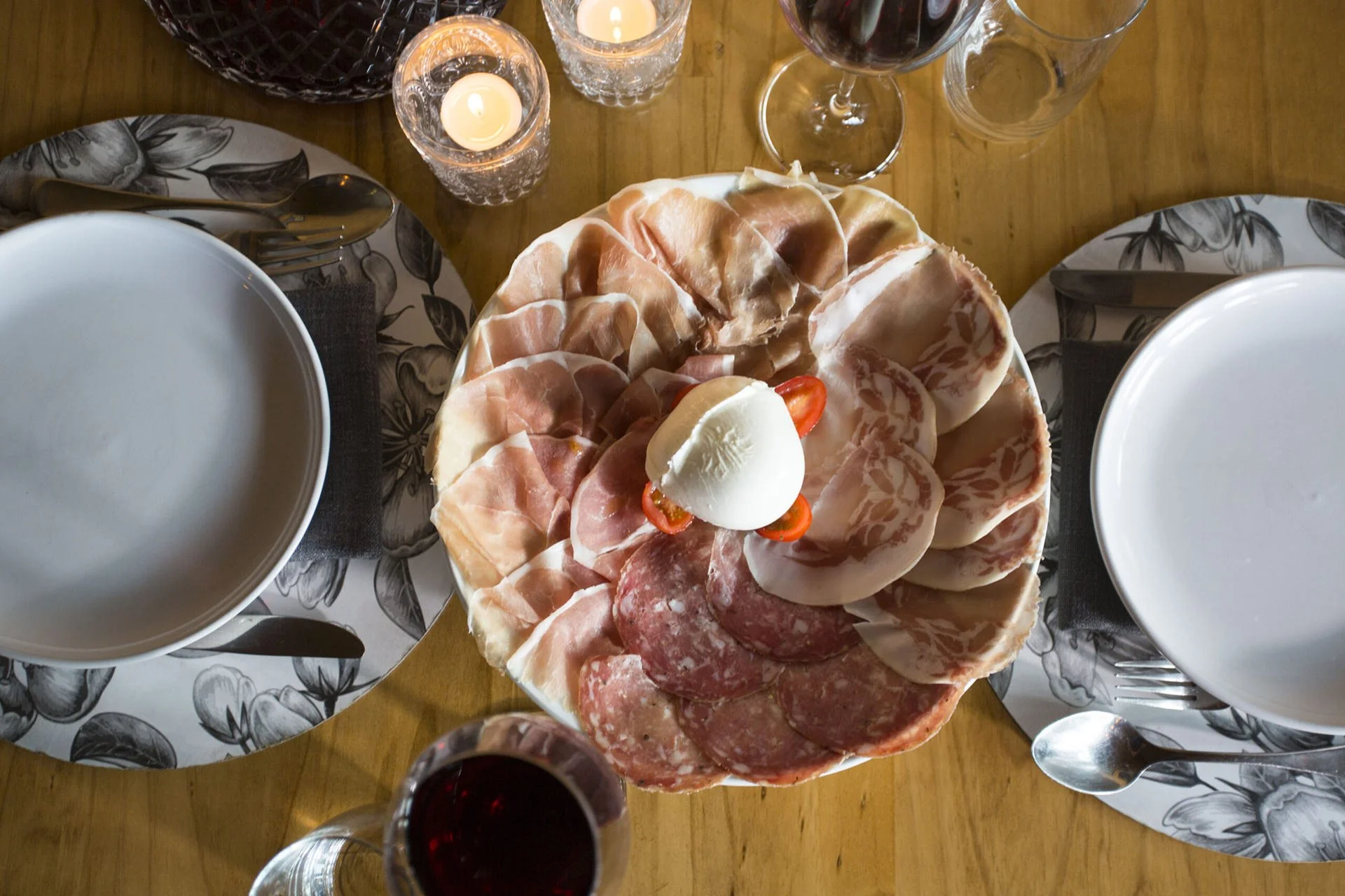 Charcuterie platter with sliced meats topped with mozzarella and cherry peppers, set on a wooden table with plates, glasses, silverware, and candles.