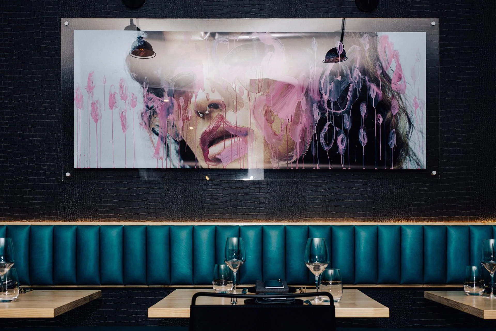 Interior of a modern restaurant or cafe with a vibrant artwork of a woman's face on the wall, black textured wall, blue padded bench seating, a wooden table set with wine glasses, and a black chair.