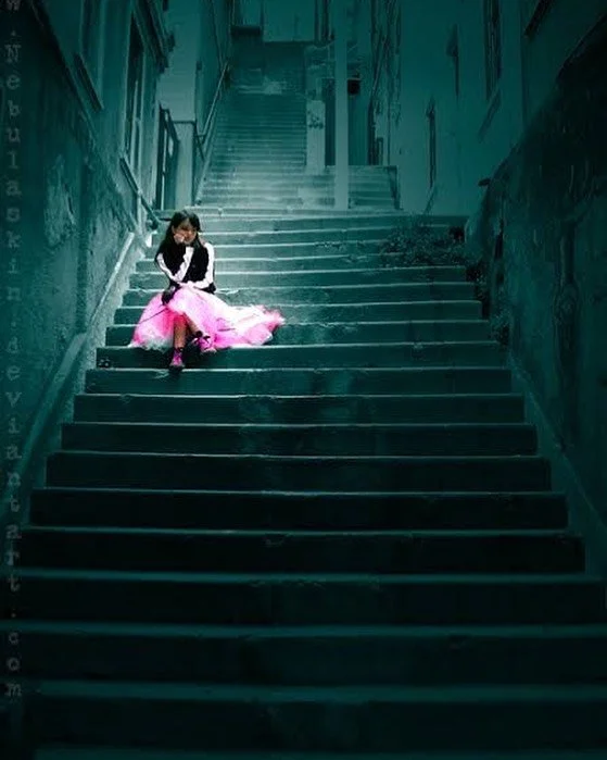 A young girl in a pink dress and black top sitting alone on a dark outdoor staircase in an alley, with graffiti on the walls.