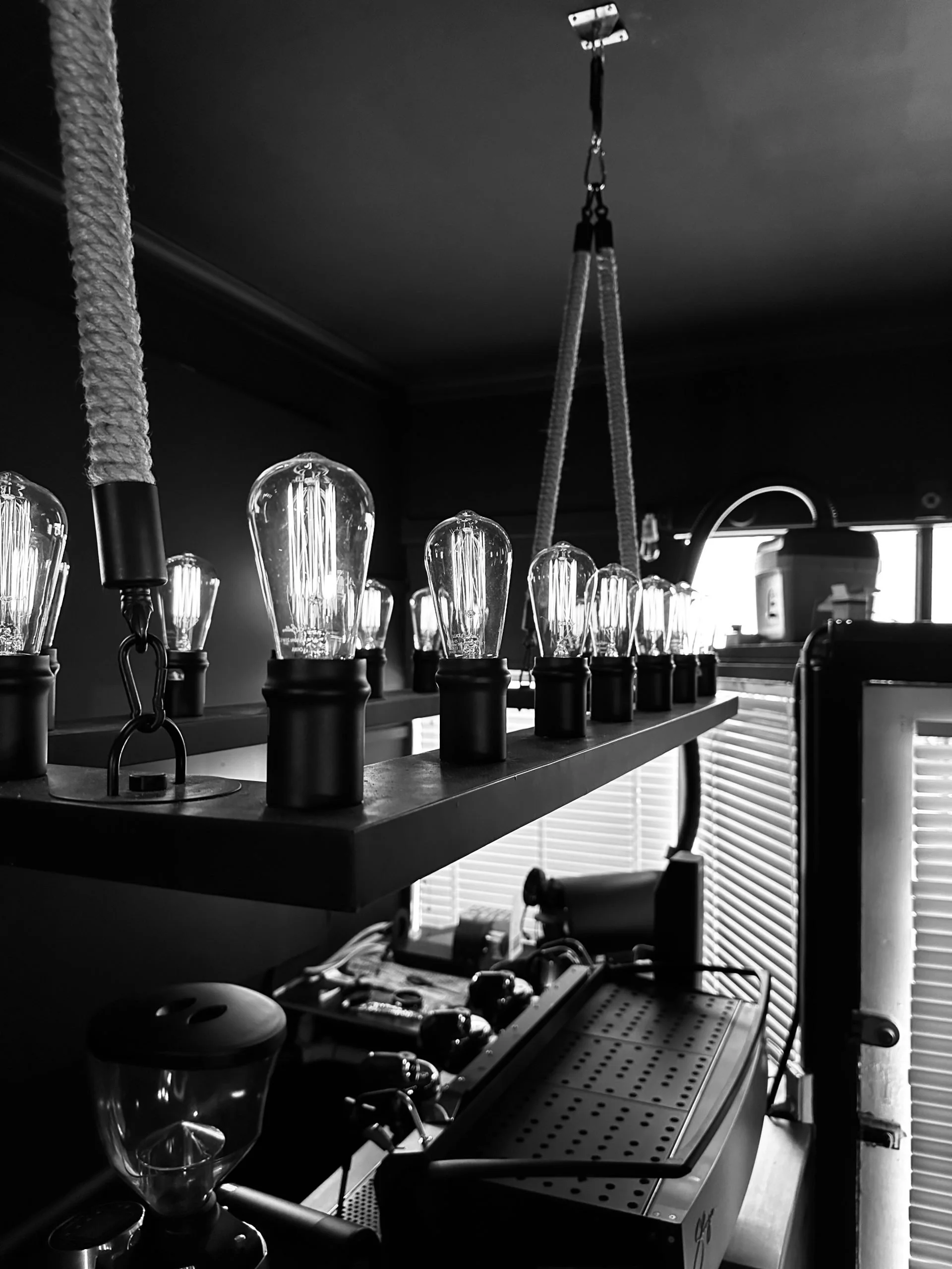 Black and white photo of an Edison-style light bulb fixture hanging from the ceiling in a coffee shop or cafe, with coffee-making equipment and a coffee grinder in the background.