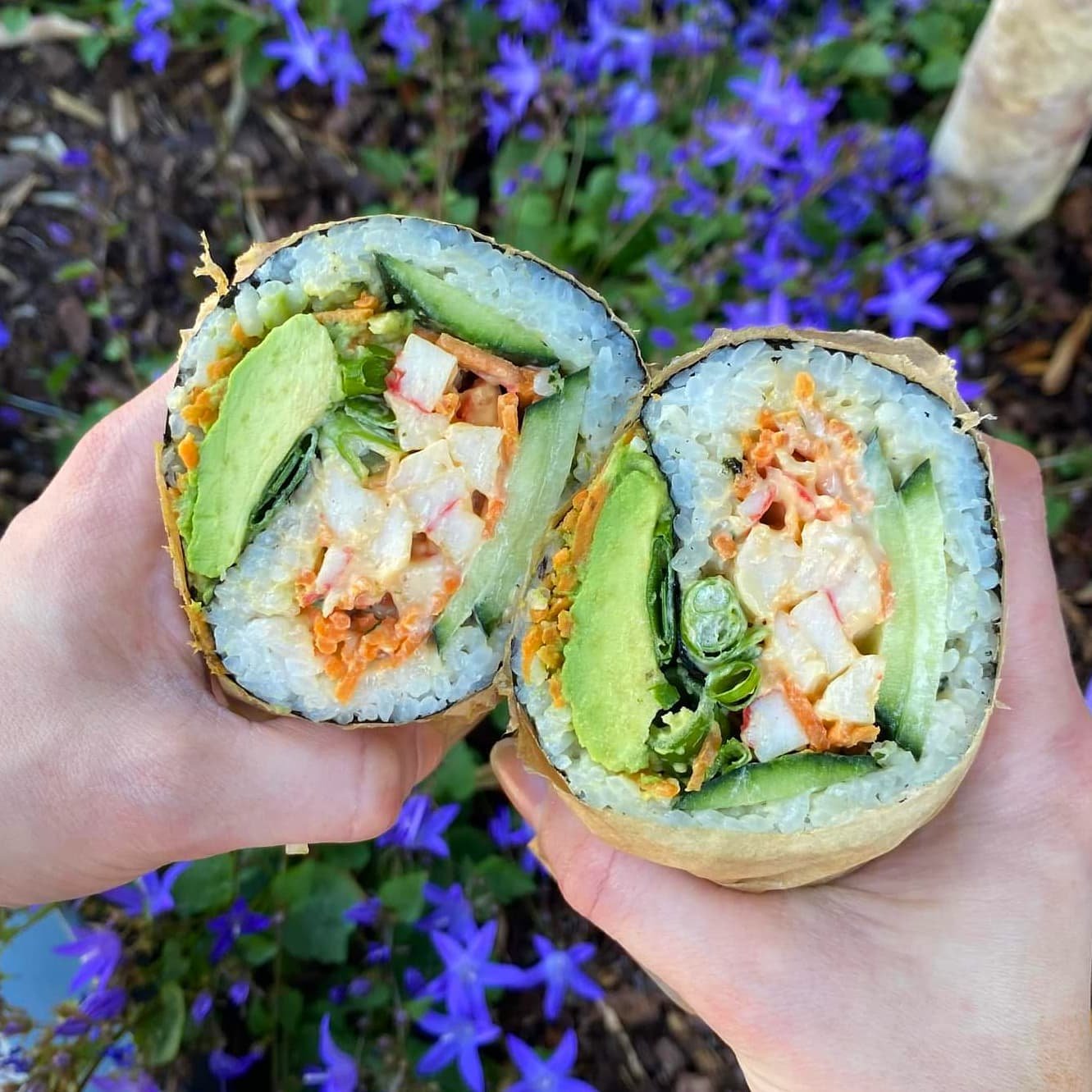 Close-up of two halves of a sushi roll with rice, avocado, cucumber, crab meat, and lettuce, held by two hands against a background of purple flowers.