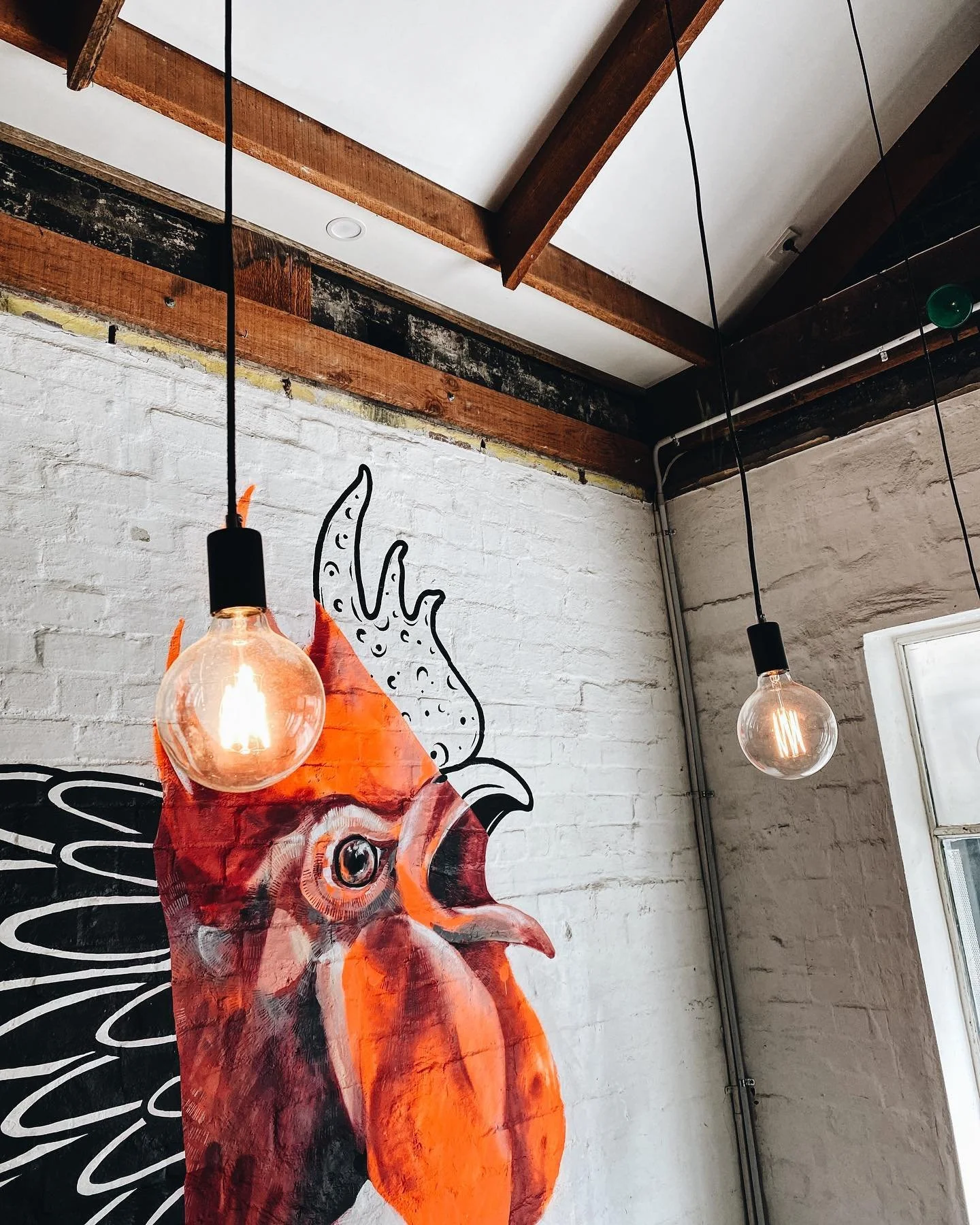 Interior of a room with white brick walls, featuring a large painted rooster on the wall. Two hanging Edison-style light bulbs with visible filaments are suspended from the ceiling with black cords.