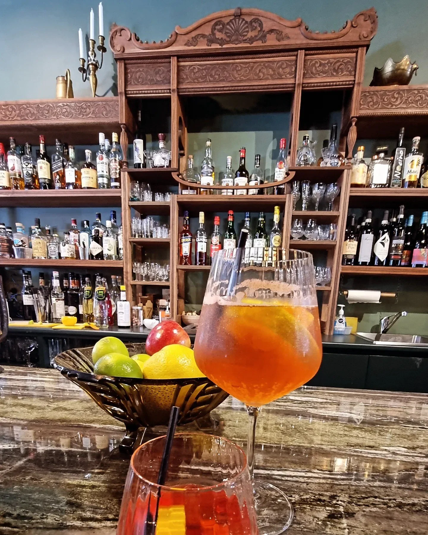 A bar with a wooden shelf filled with various bottles of alcohol and glassware. In the foreground, there is a bowl of apples and lemons, and two cocktails on the bar counter, one red and one orange, with black straws.