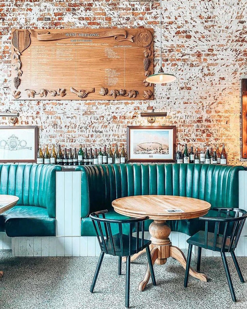 Interior of a restaurant with a green booth, a round wooden table, and black chairs. A brick wall with wine bottles, framed pictures, and a wooden menu board with grapevine decorations. Hanging light fixture.