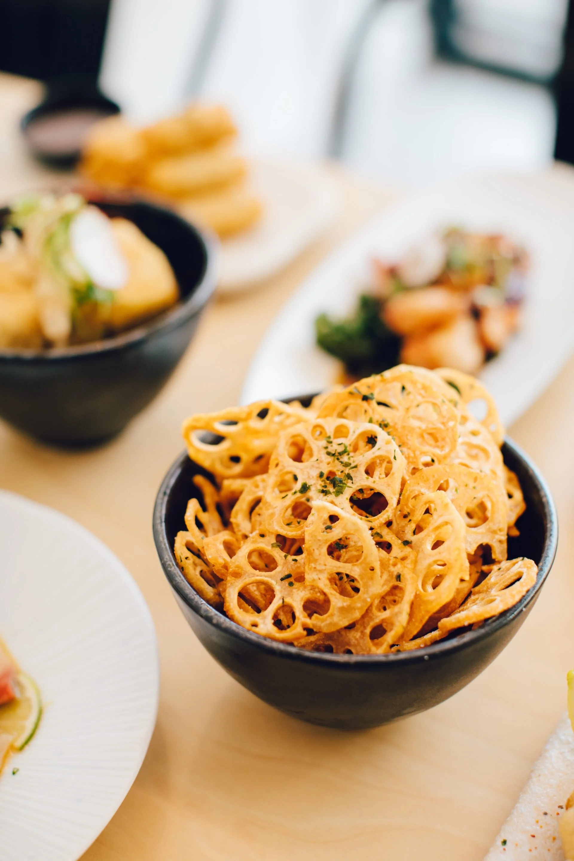 A black bowl filled with crispy lotus root chips garnished with herbs, placed on a light-colored table with various Asian dishes in the background.