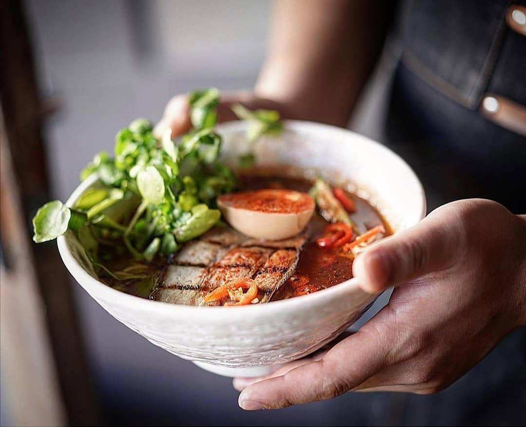A person holding a bowl of Asian-style noodle soup with fish, boiled egg, vegetables, and broth.