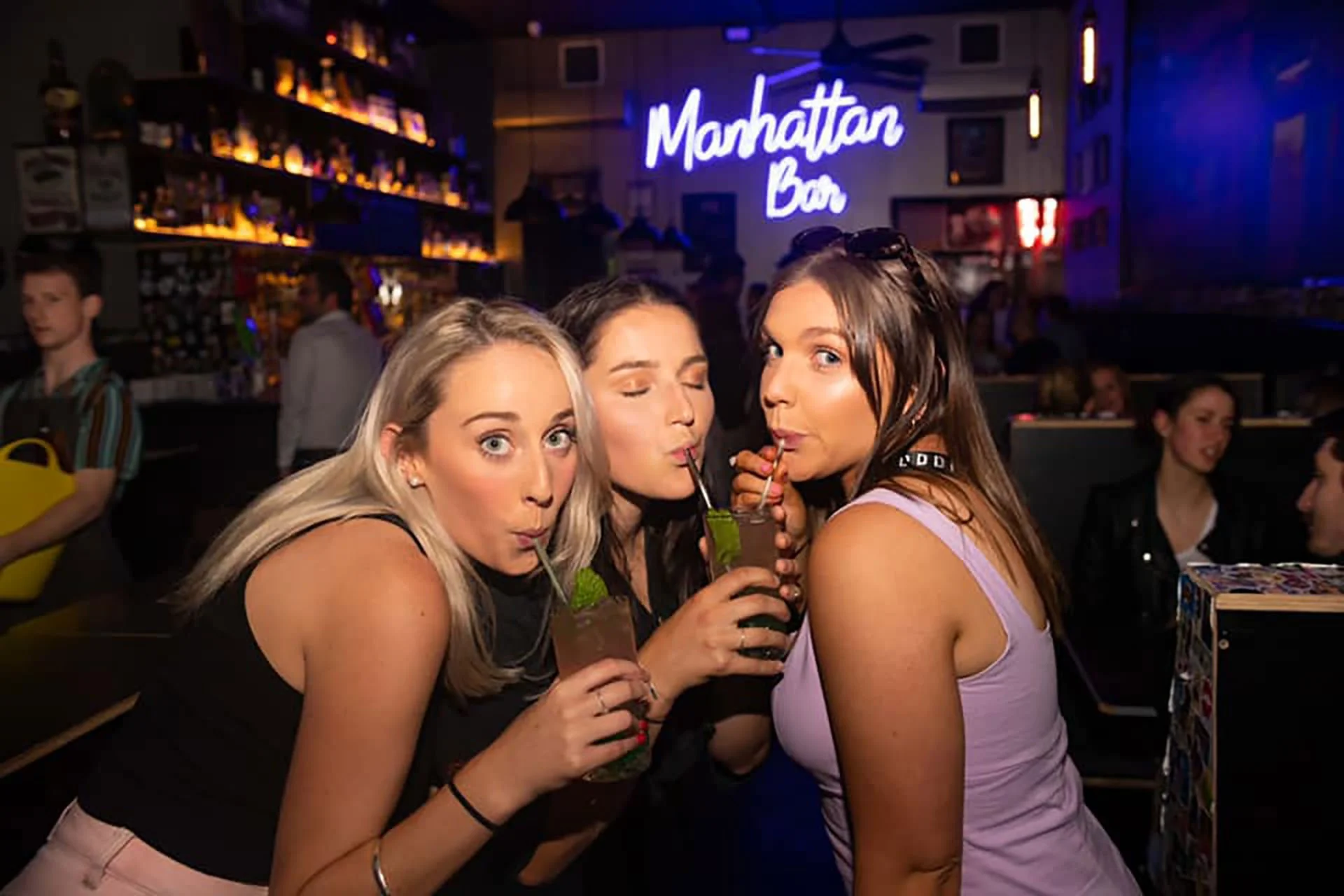 Three women enjoying drinks at Manhattan Bar, celebrating and drinking cocktails with lime garnish, in a lively bar setting with a neon sign and other patrons in the background.