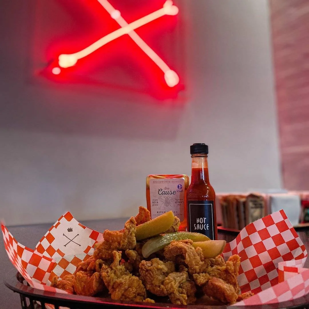Basket of fried chicken pieces with pickles, hot sauce, and honey in a restaurant setting, illuminated by a red neon clock on the wall.