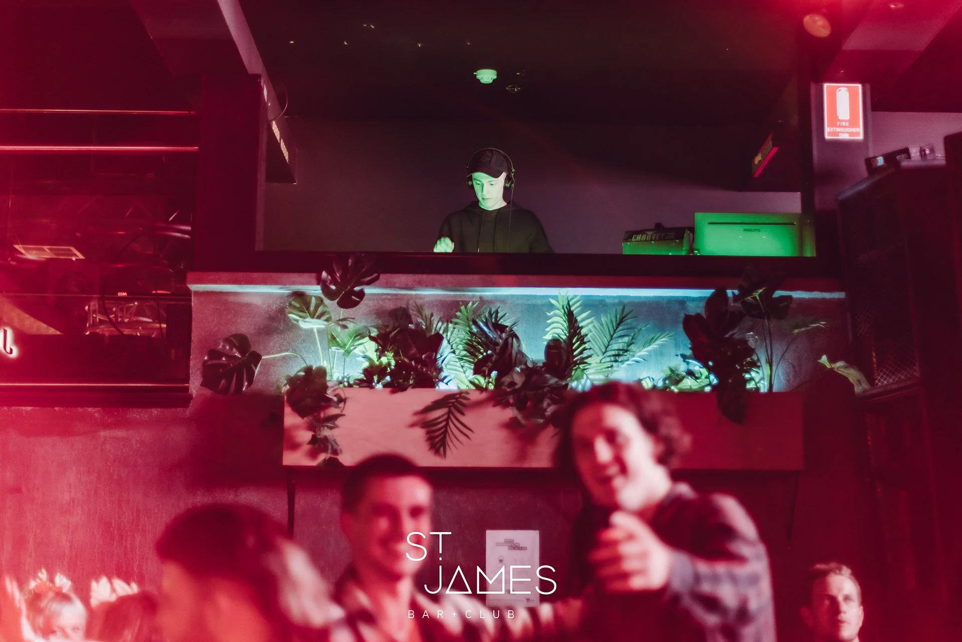 A DJ wears headphones and a black shirt, standing behind a DJ booth with plants, in a nightclub. The club has pink and green lighting, and people are dancing and socializing in the foreground.