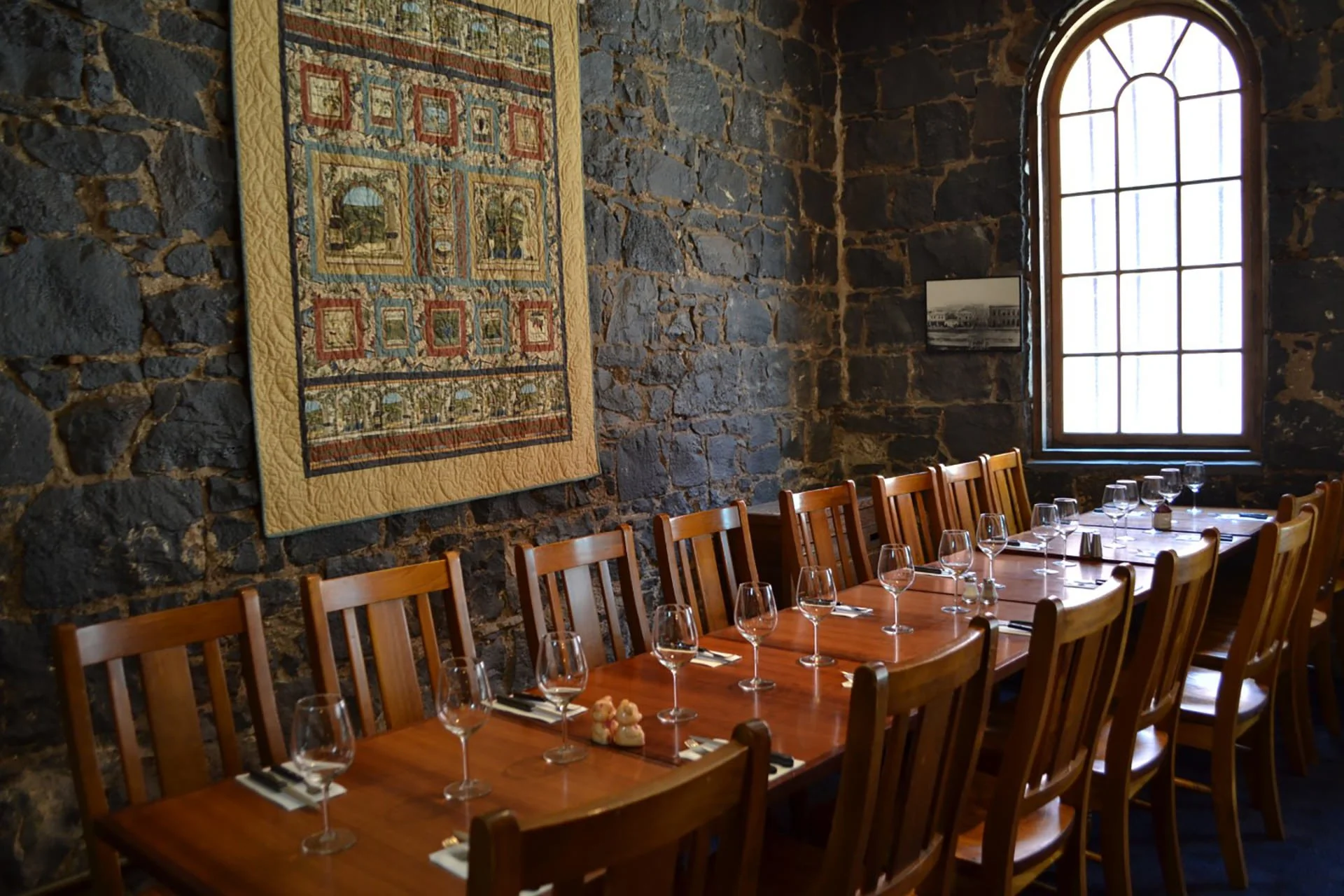 A dining room with a long wooden table set with wine glasses, utensils, and salt and pepper shakers. The room has a stone wall, a quilt hanging on the wall, and a large arched window.