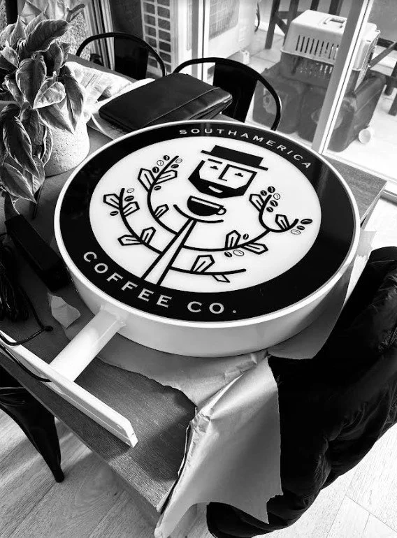 A large round sign resembling a coffee cup lid with the logo of South America Coffee Co., featuring a smiling man with a hat and a beard, surrounded by leaves. The sign is placed on a table in a cafe setting.