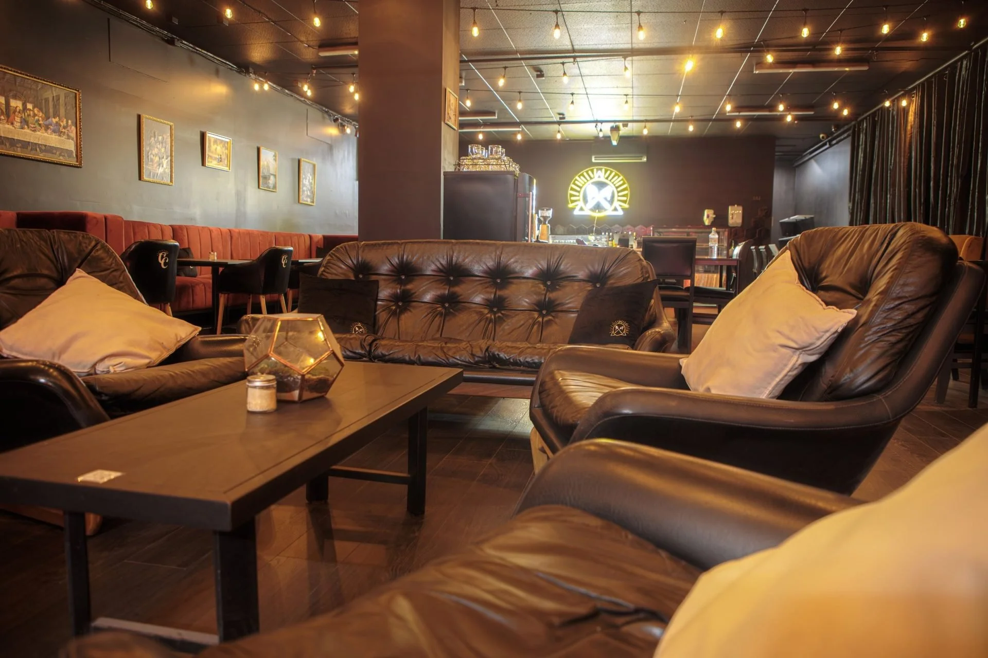 Cozy lounge with leather sofas, pillows, a wooden coffee table with a candle and a geometric decor piece, and a bar in the background with warm lighting and a neon sign.