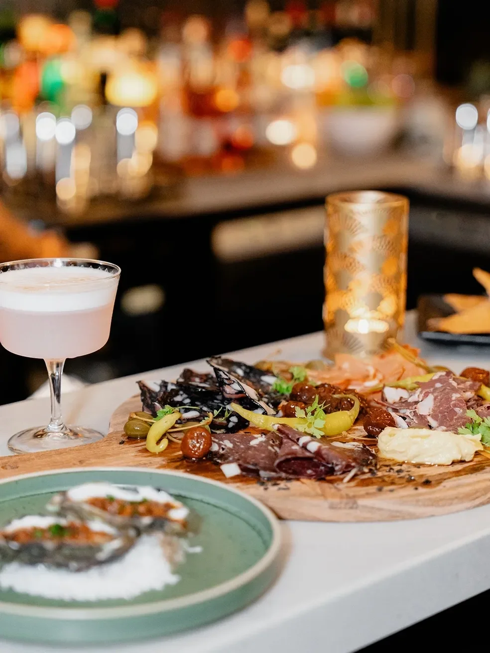 A charcuterie board with various meats, cheeses, olives, and pickles, a pink cocktail in a coupe glass, and a small dish with oysters, set on a table with a candle and blurred background of a bar or restaurant ambiance.