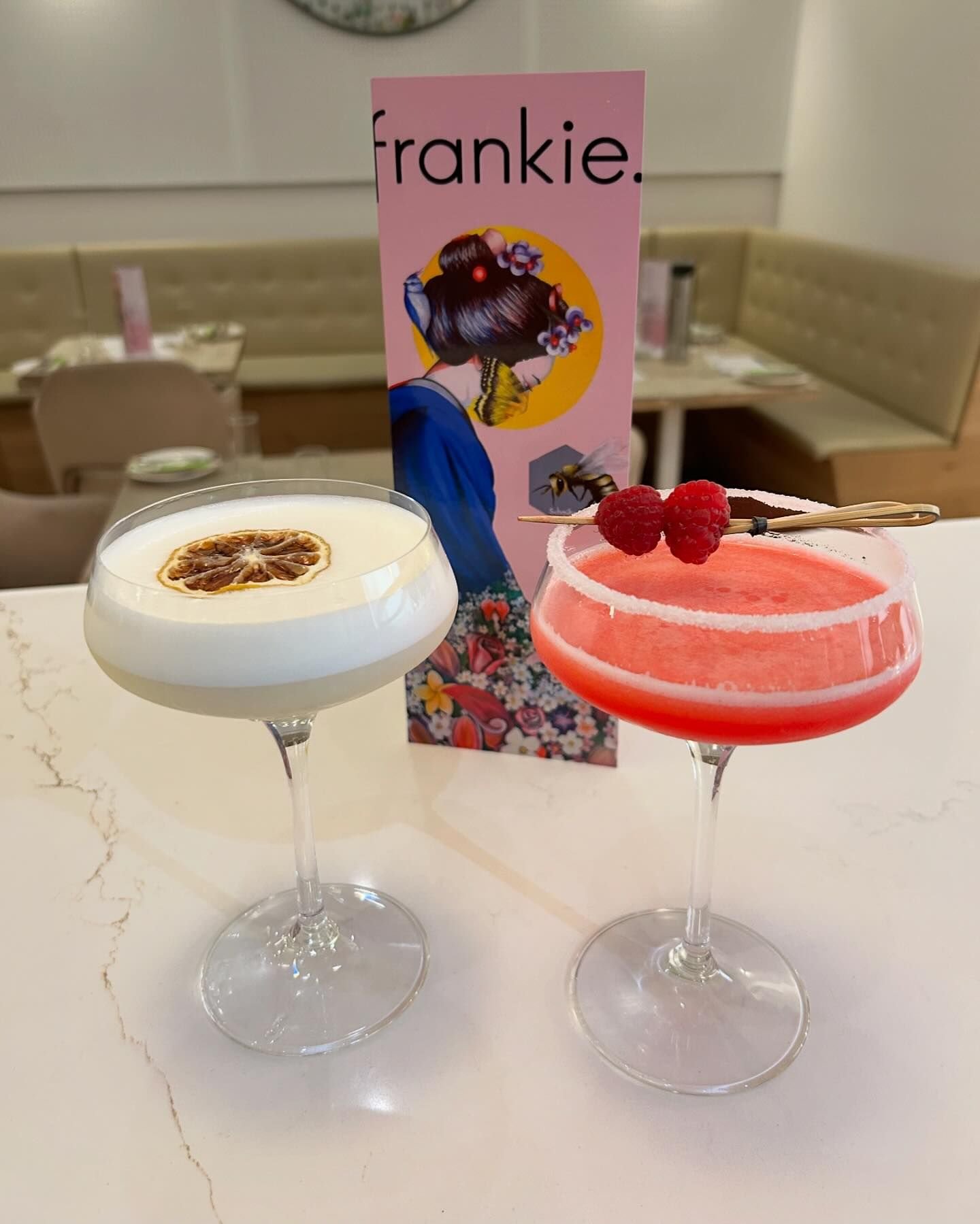 Two cocktails on a white table, one with a lemon slice garnish and the other with raspberries, with a pink menu featuring a woman in traditional attire and a bee in the background.