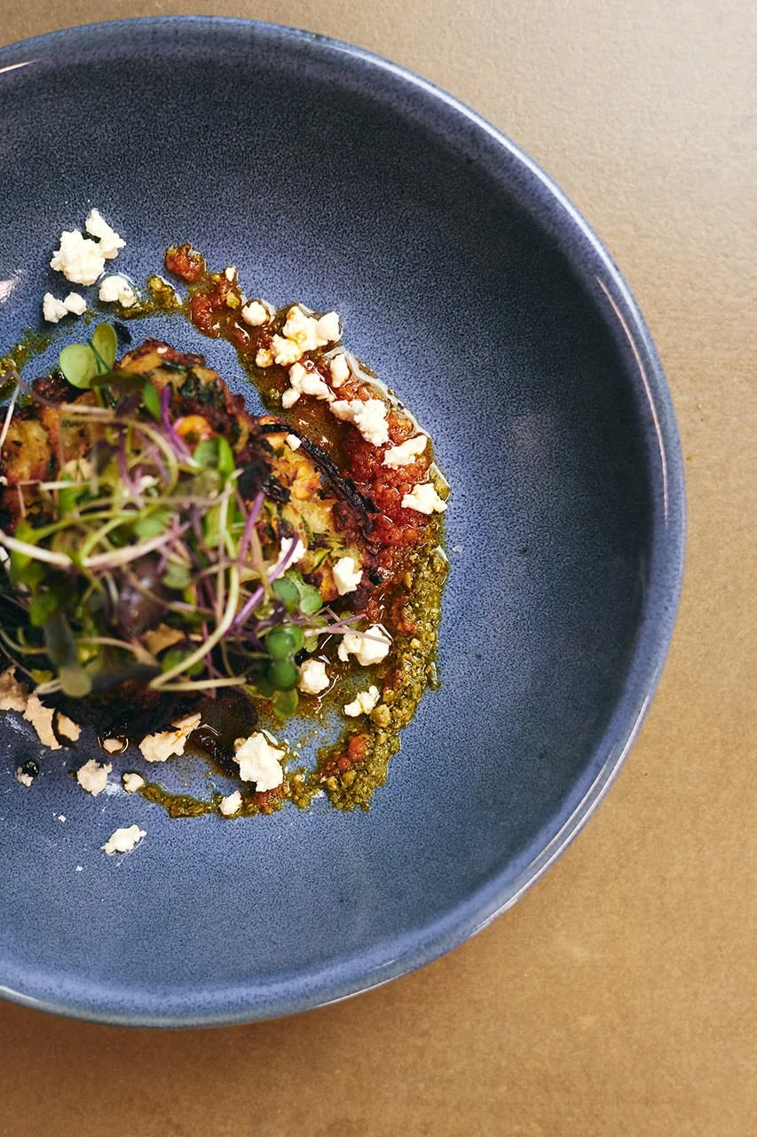 Close-up of a plated dish featuring grilled vegetables topped with microgreens, crumbled cheese, and a drizzle of sauce on a dark blue plate.