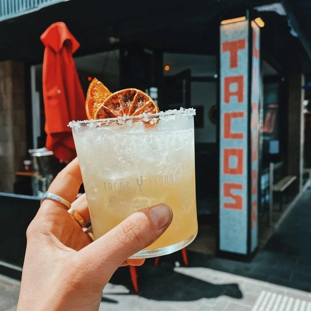 Hand holding a cocktail garnished with dried orange slices in front of a taco shop with a vertical sign that reads 'TACOS' outside.