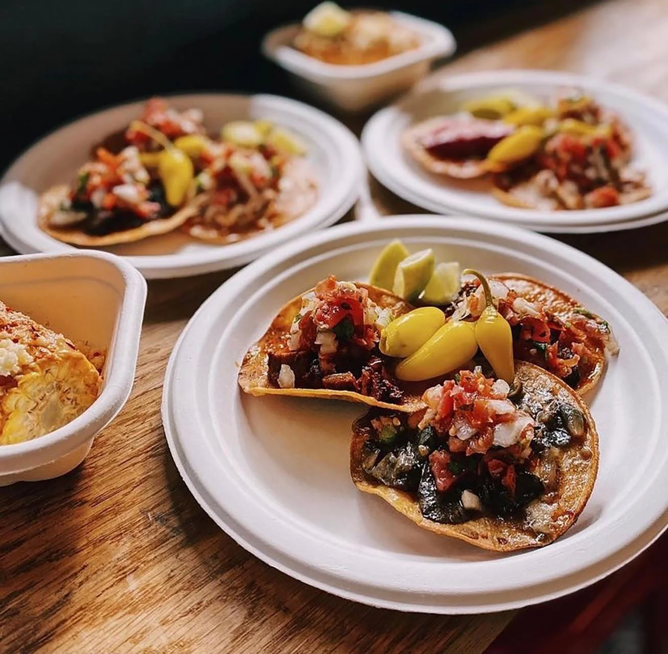 Tacos filled with meat and topped with pickled peppers and chopped onions, served on white plates with small bowls of salsa on the side.