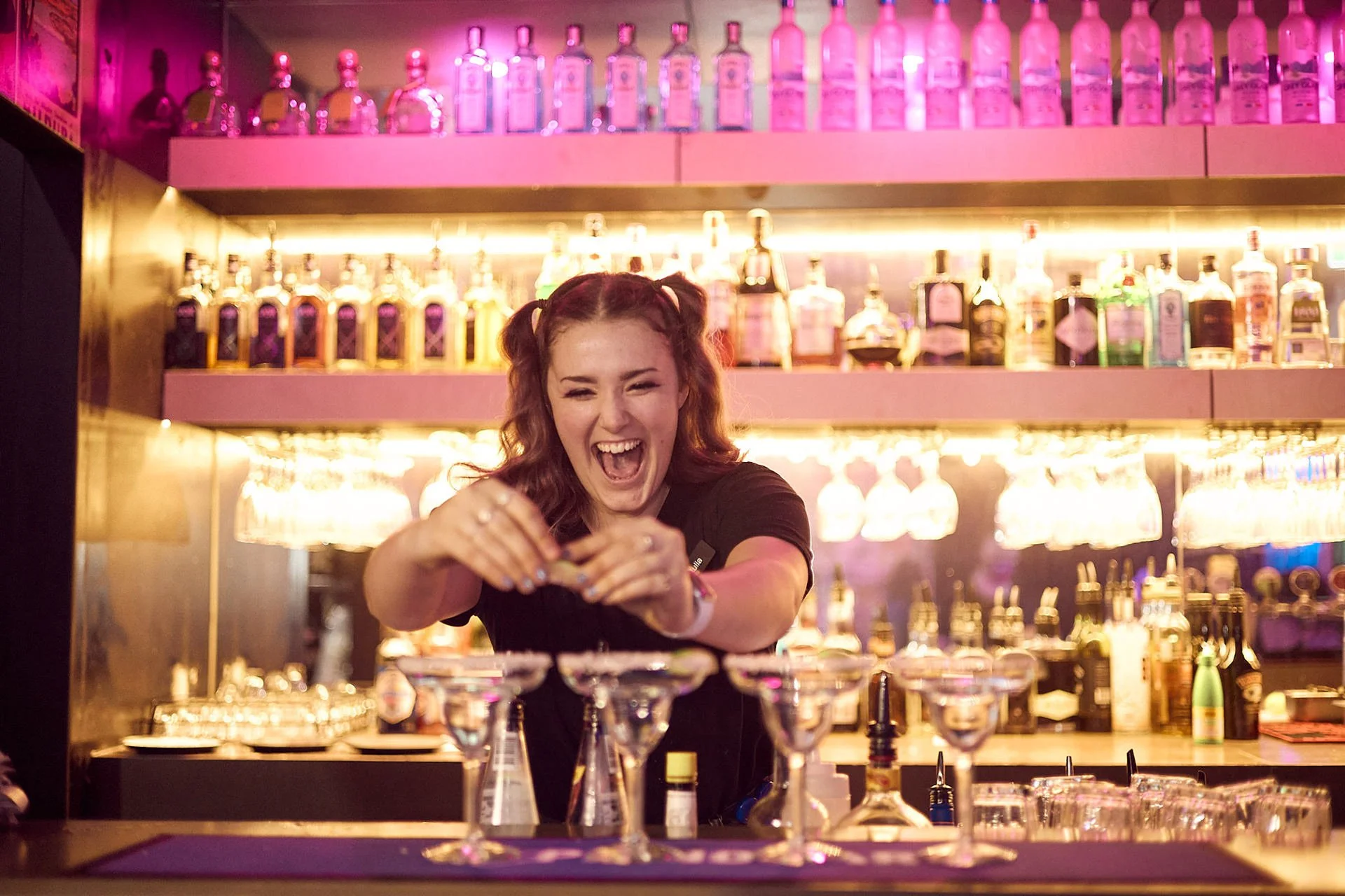 A bartender with red hair styled in pigtails laughing joyfully while preparing cocktails at a bar with a colorful, illuminated backdrop of liquor bottles.