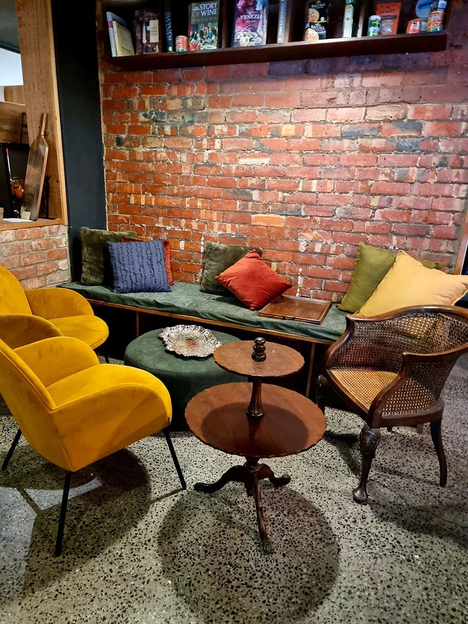 Cozy seating area with two yellow velvet armchairs, a green velvet bench with multiple colorful cushions, a rattan armchair, a small round wooden table, a green ottoman with a silver decorative tray, brick wall background, and a bookshelf above with 