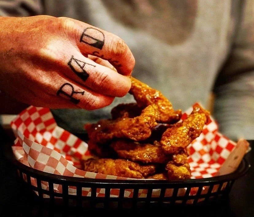 A hand with tattoos on fingers reaching into a basket of fried chicken wings lined with red and white checkered paper.