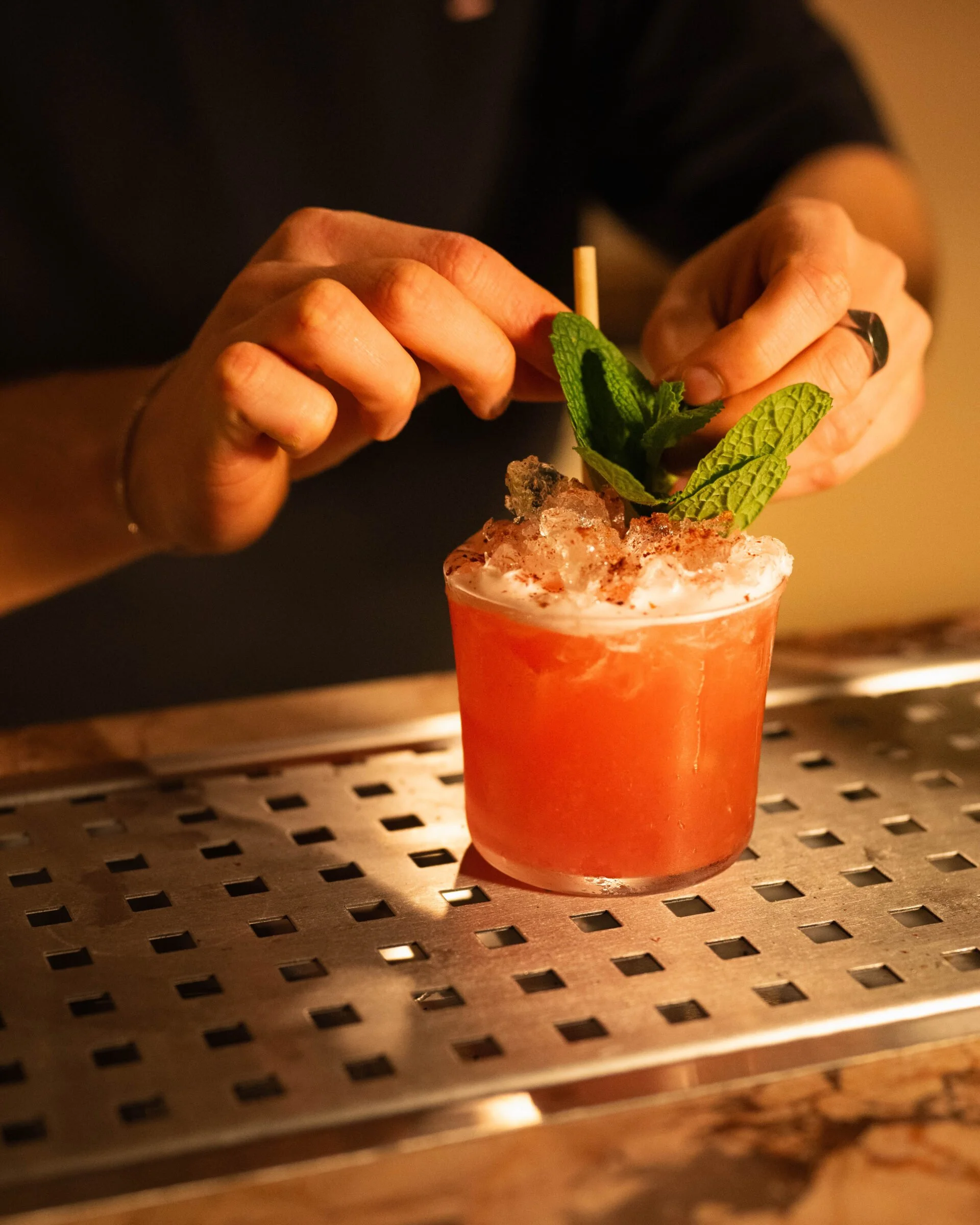 A bartender garnishing a red cocktail with mint leaves and a stick, on a metallic surface.