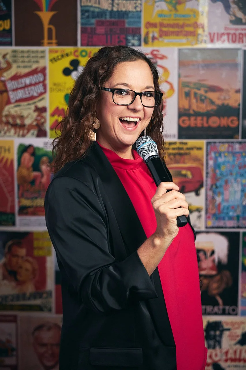A woman with wavy brown hair, glasses, and earrings, holding a microphone, smiling, wearing a black jacket over a red top, standing in front of a wall covered with colorful vintage posters.