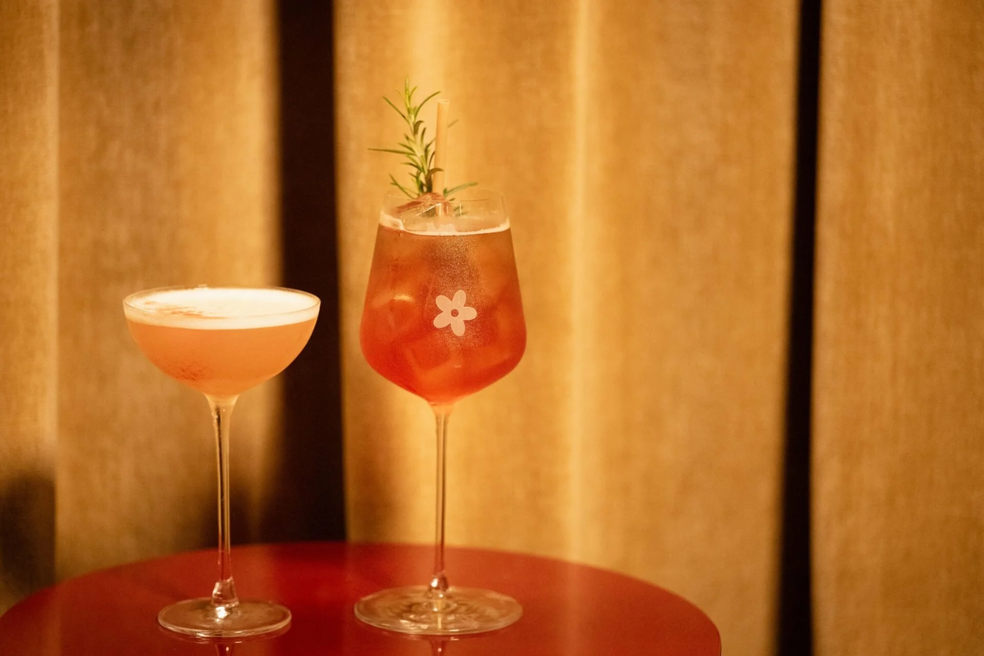 Two cocktails on a wooden table against a beige curtain background. The left cocktail is in a coupe glass with light pink liquid. The right cocktail is in a tall glass with pinkish drink, garnished with a sprig of rosemary and ice cubes, with a white