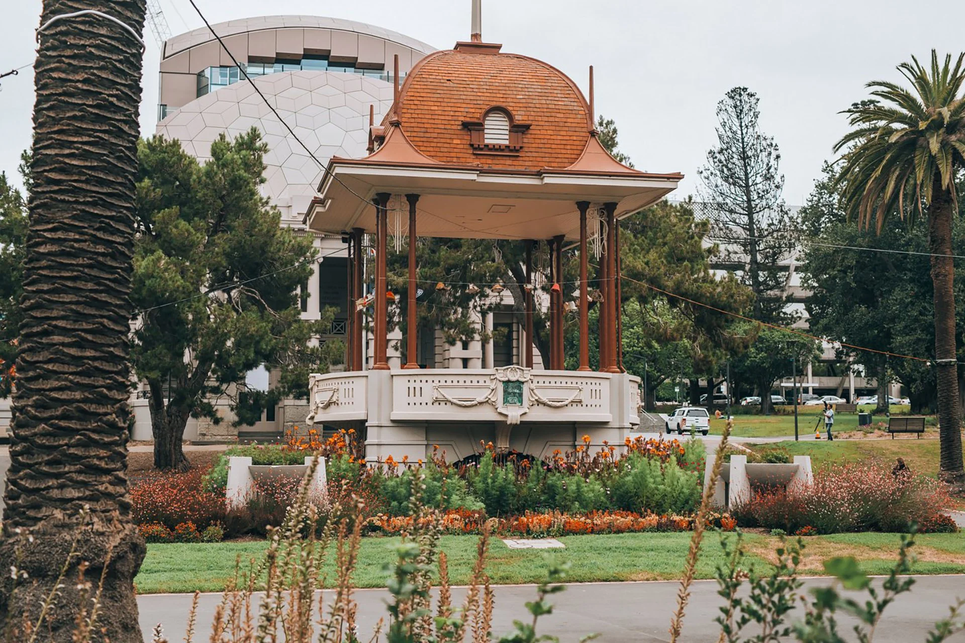 A Victorian-style bandstand with a red-tiled roof in a park, surrounded by flowers, trees, and a cityscape in the background.