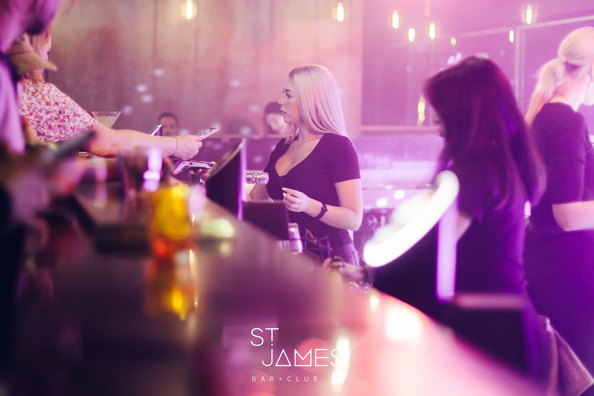 A young woman in a black top leaning on a bar counter in a nightclub with neon lights and other people. The text 'ST JAMES BAR + CLUB' is visible at the bottom of the image.