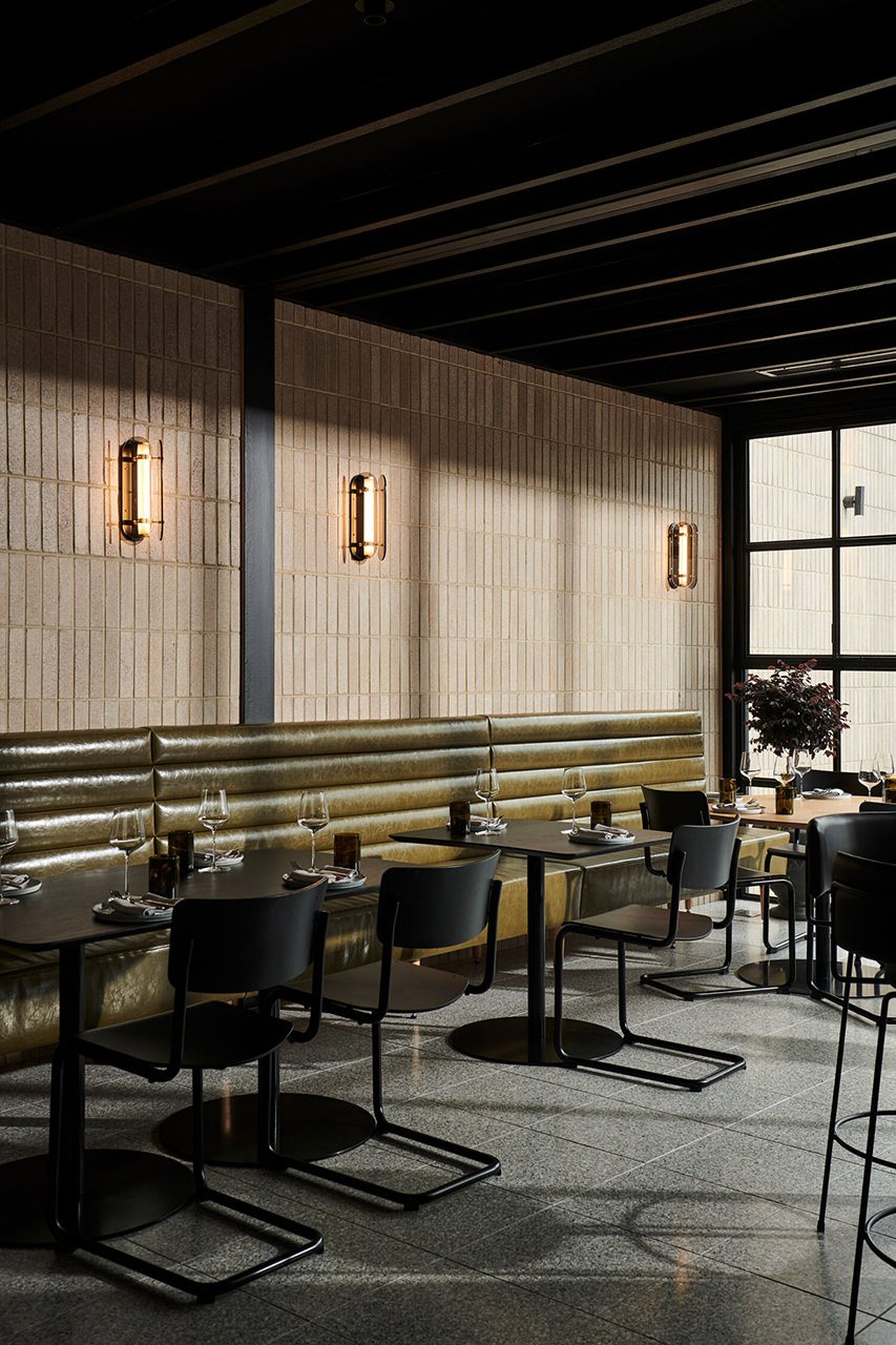 Interior of a modern restaurant with black tables, black chairs, and a long gold upholstered banquette against a textured beige wall, bathed in warm lighting from wall sconces.